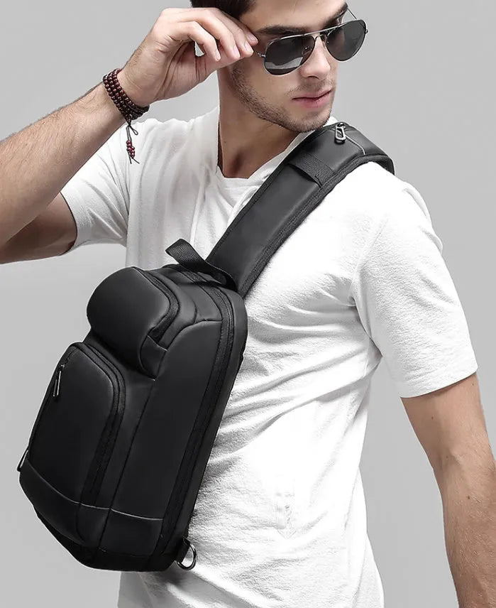 Black square-shaped outdoor shoulder bag with a single adjustable strap, shown with open zipper revealing internal laptop and phone pockets.