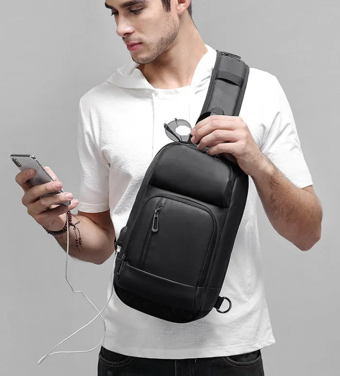 Black square-shaped outdoor shoulder bag with a single adjustable strap, shown with open zipper revealing internal laptop and phone pockets.