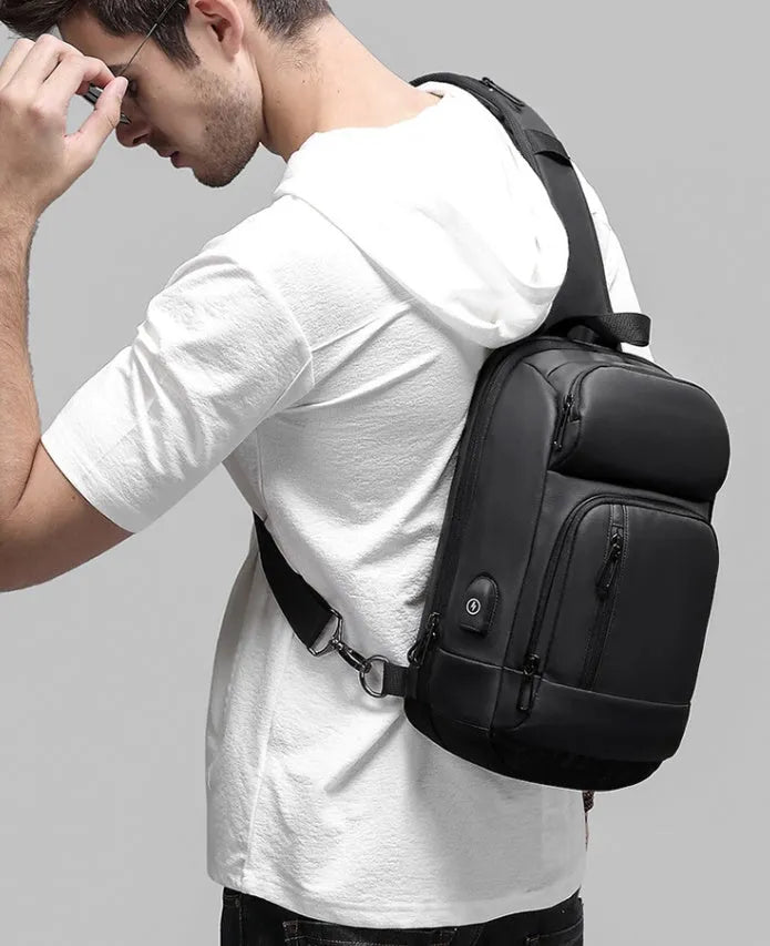 Black square-shaped outdoor shoulder bag with a single adjustable strap, shown with open zipper revealing internal laptop and phone pockets.