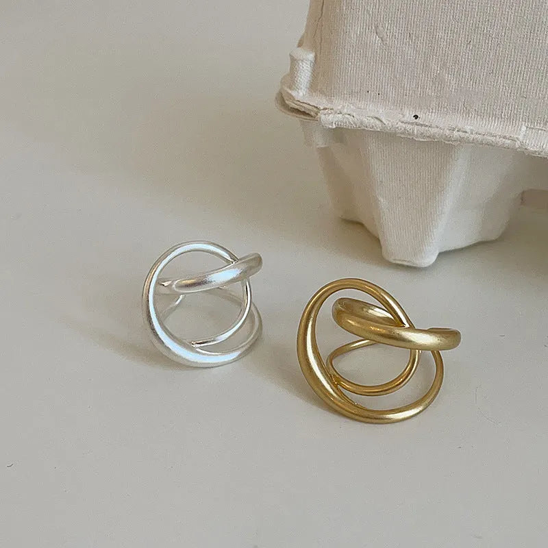 Geometric open ring for women with a 3D interlaced design in matte metal, shown in gold and silver finishes.