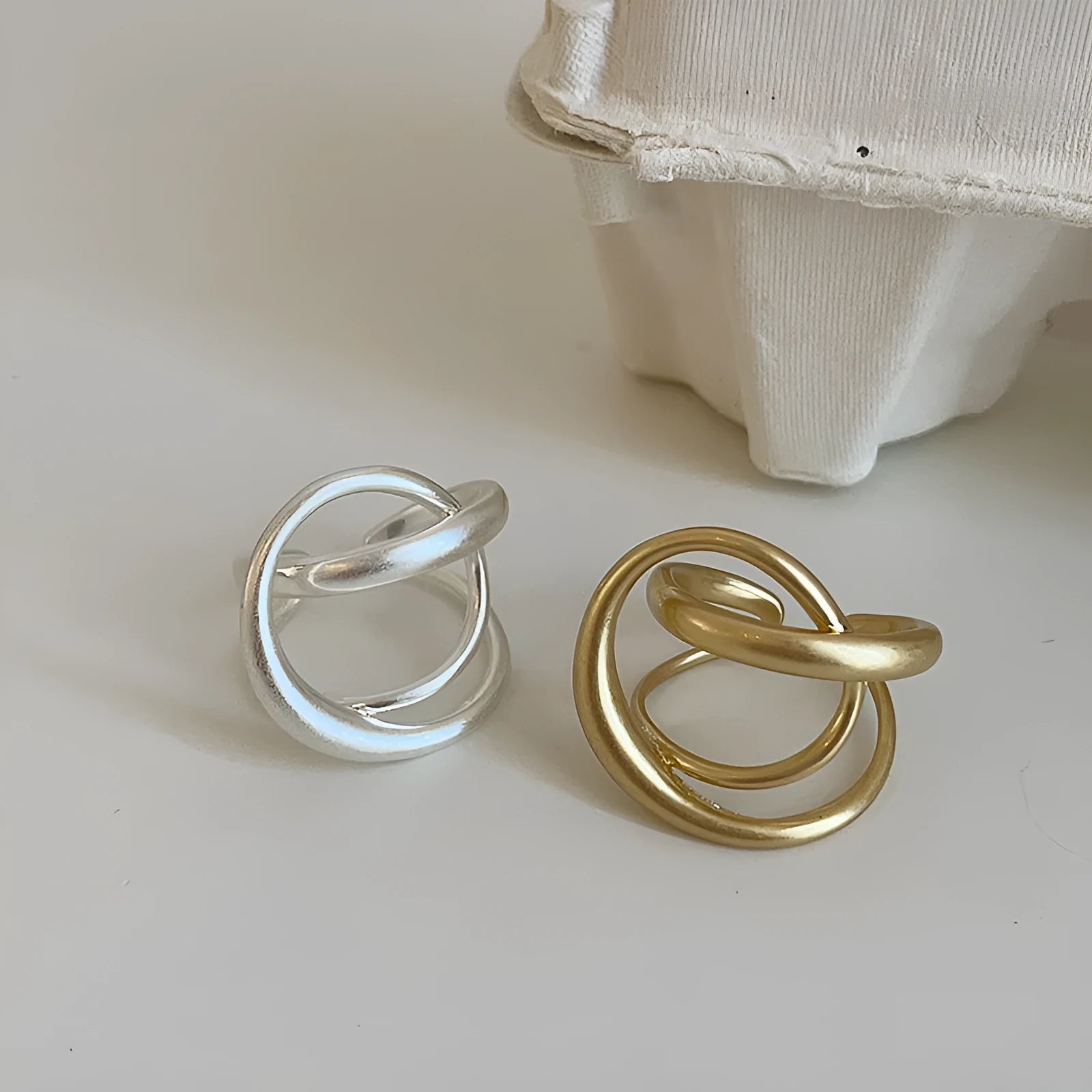 Geometric open ring for women with a 3D interlaced design in matte metal, shown in gold and silver finishes.