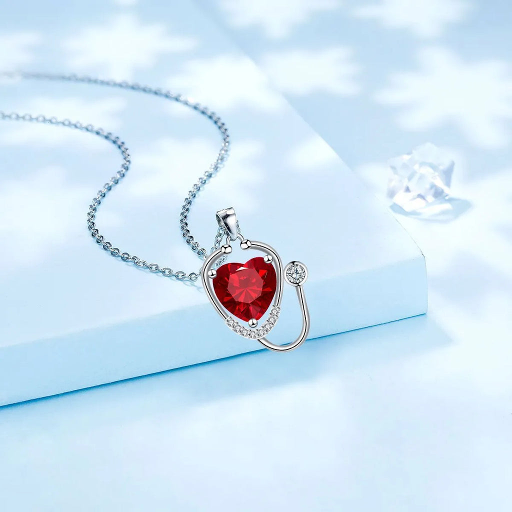 Red diamond stethoscope necklace with a wing and cross pendant on a chain.