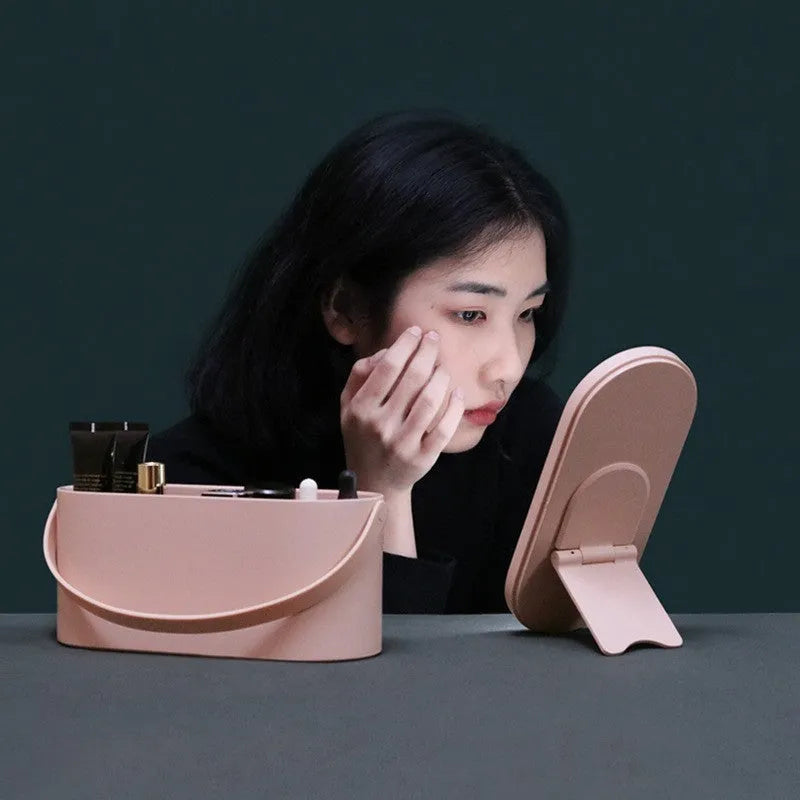 Portable LED light storage box beauty and makeup mirror with aluminium mirror lens in pink colour on a bedroom surface.