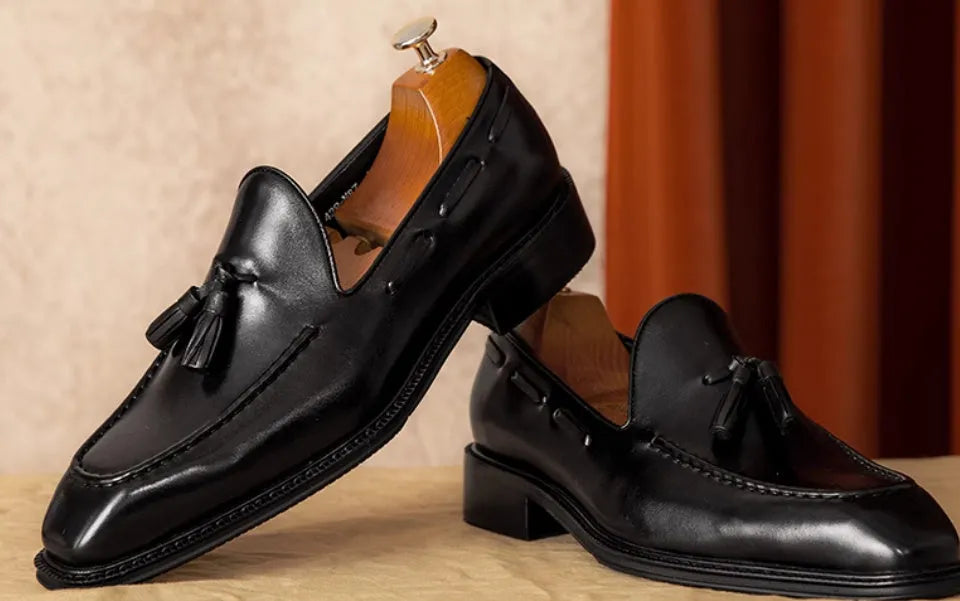 Men's black leather slip-on loafers with a fringed detail on the vamp and a rubber sole.