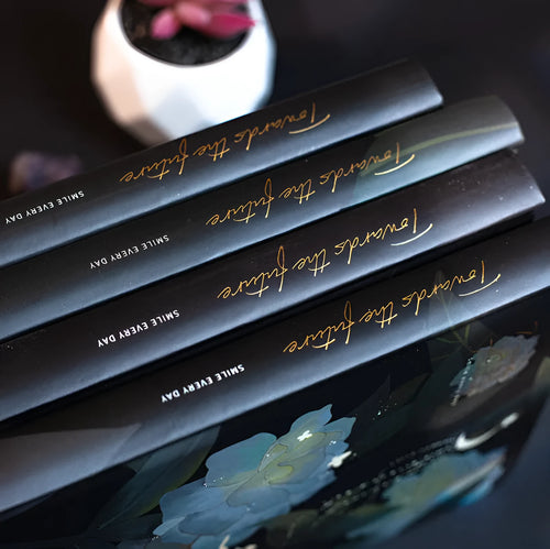A5 notebook with luminous reflective cover shown in four colours: deep blue, gold, light blue, purple, open to reveal pages with colour illustrations.